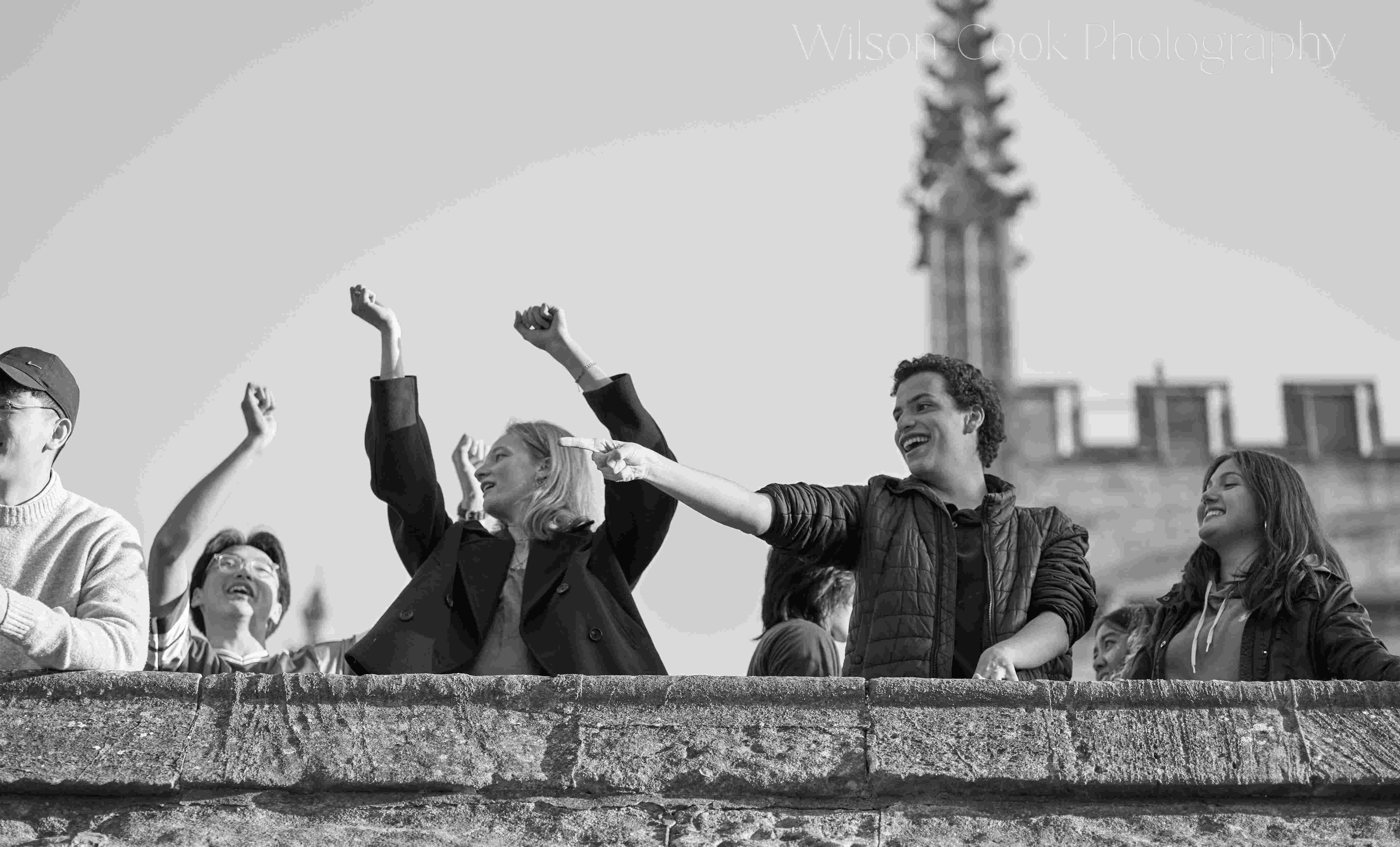 People cheering, viewed from below, it looks as though they are on the top of a historic building. The figure on the right is pointing at something they have just noticed.
