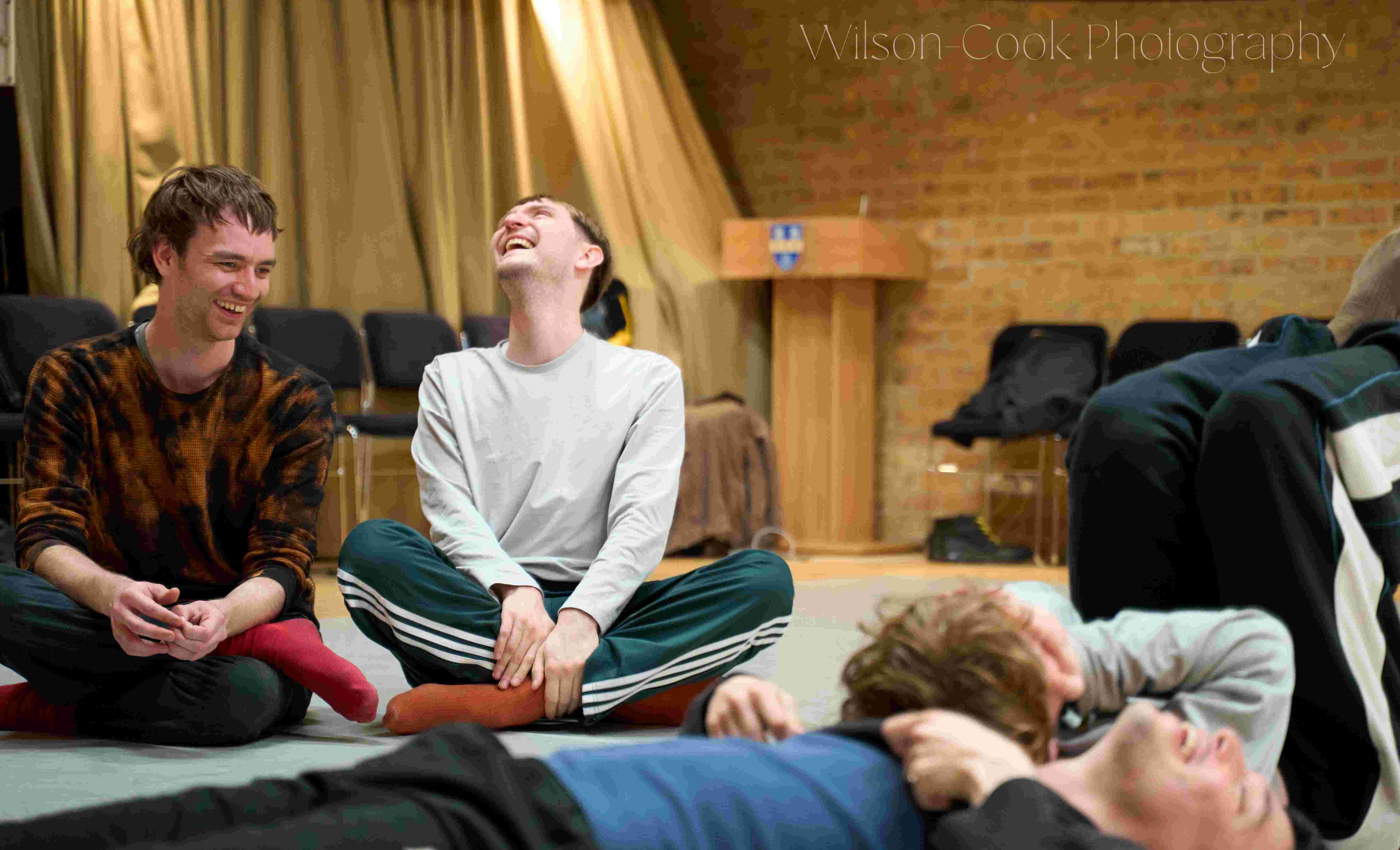 Four people laughing, two of them are laying on the floor, the other two are sitting cross-legged nearby.