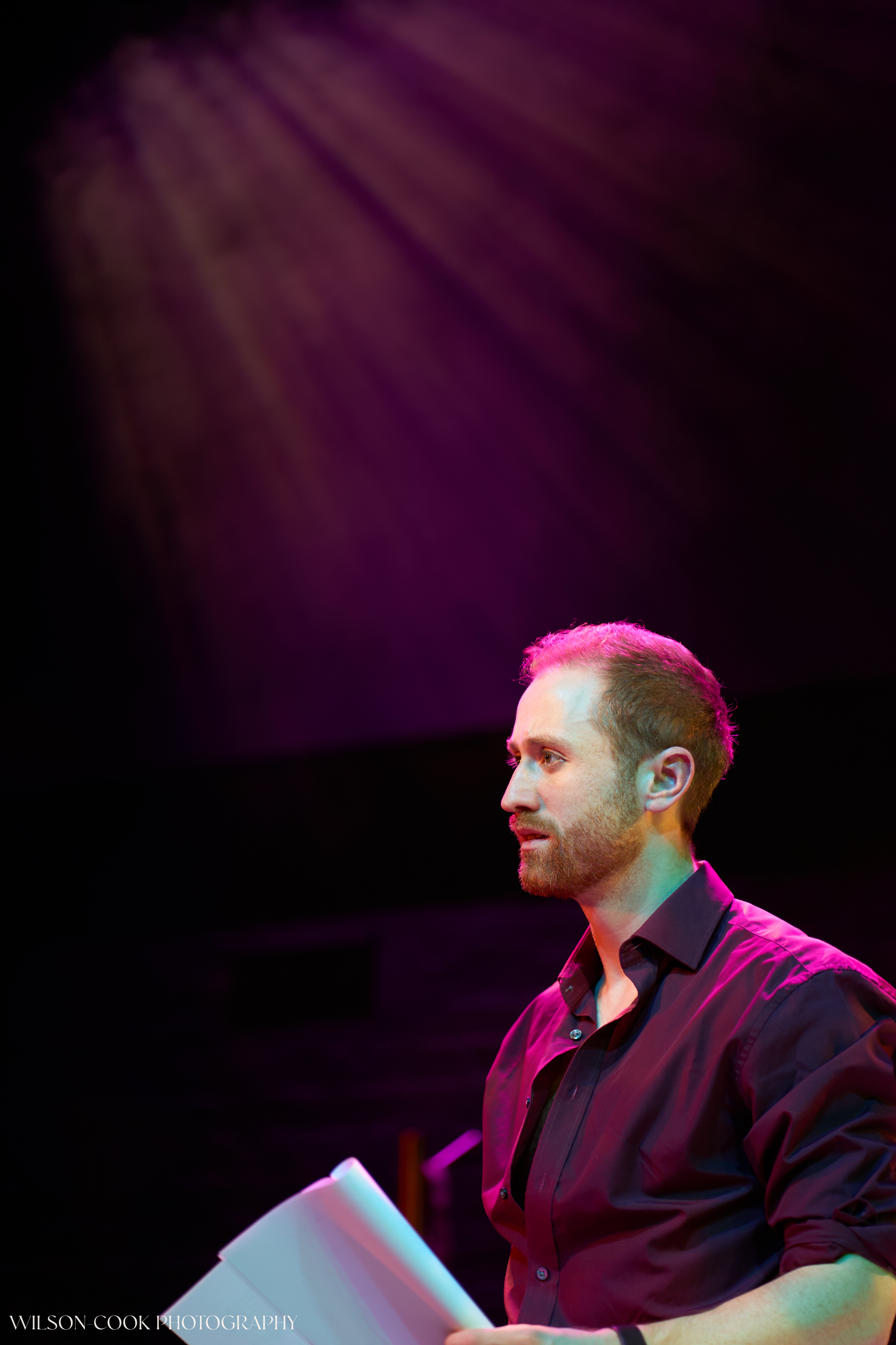 2025.  A man on stage with dramatic purple lighting behind him, he is holding what looks like a script and is mid-speech.