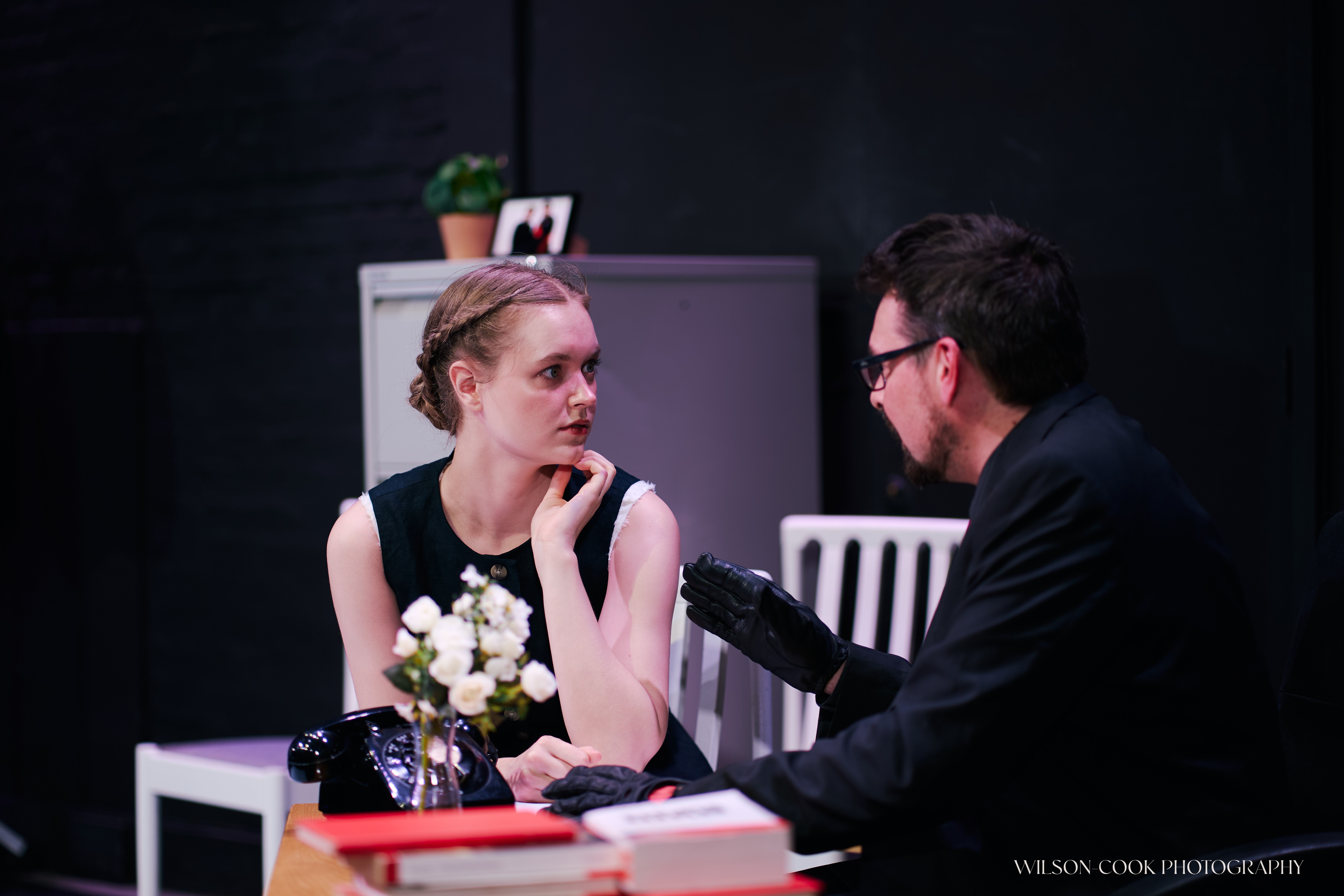 2025. Two actors on stage at a table - a woman is looking concerned at a man to her left.