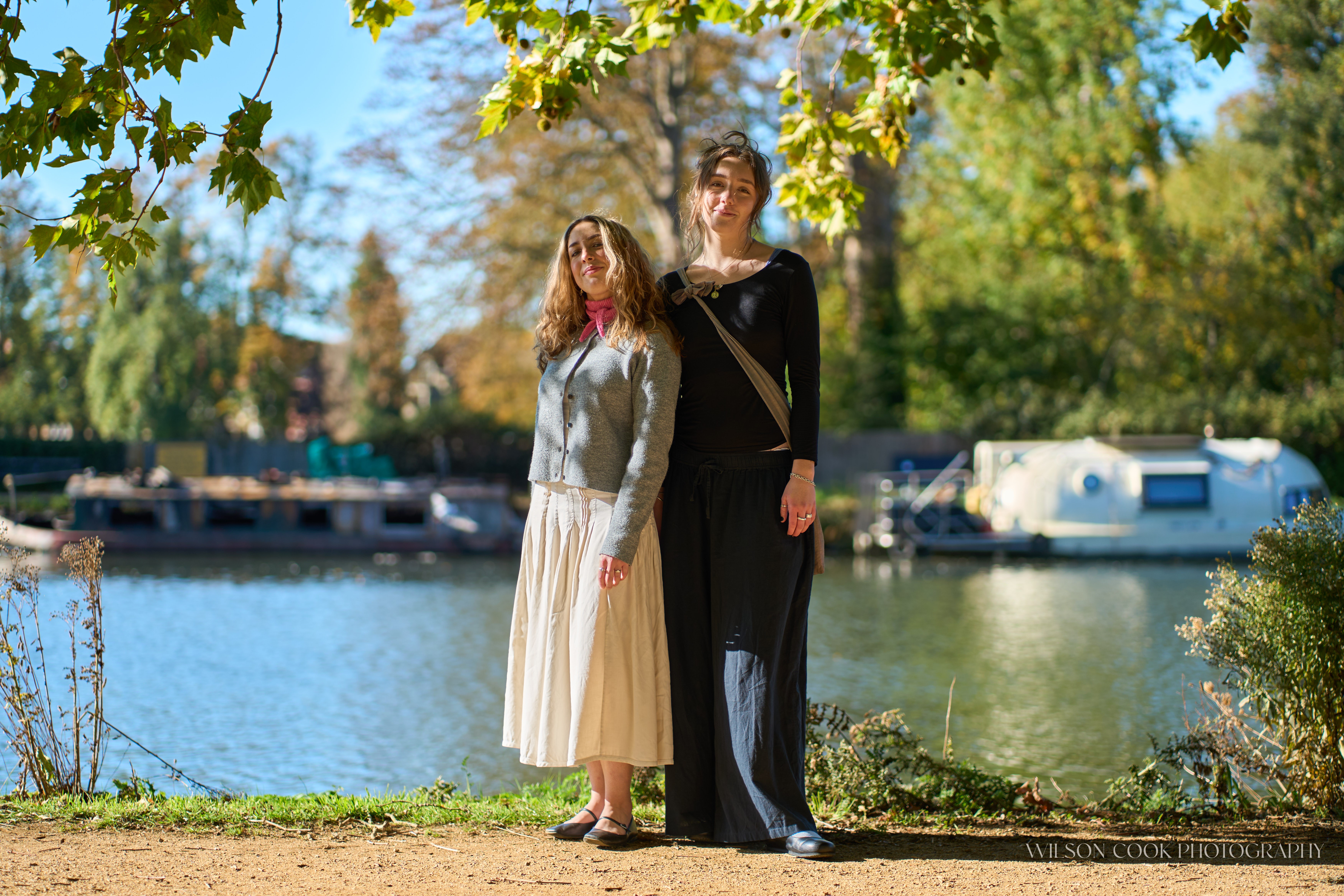 2025.  Two women standing in front of the river, they are under a tree and the dappled light is casting shadows across them.
