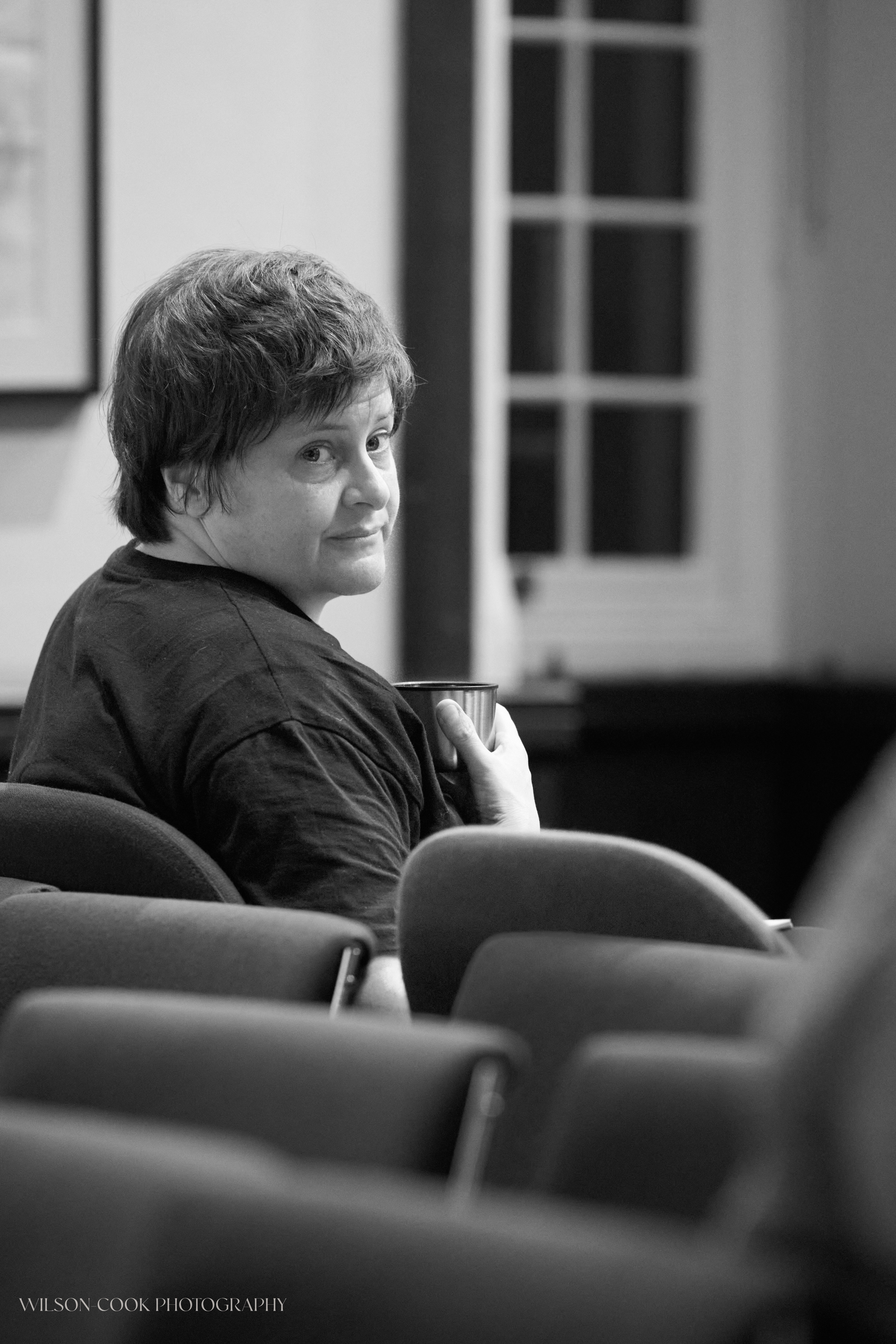 2025.  A black and white image of a woman, she is holding a thermos flask and looking over her right shoulder seemingly towards someone else.