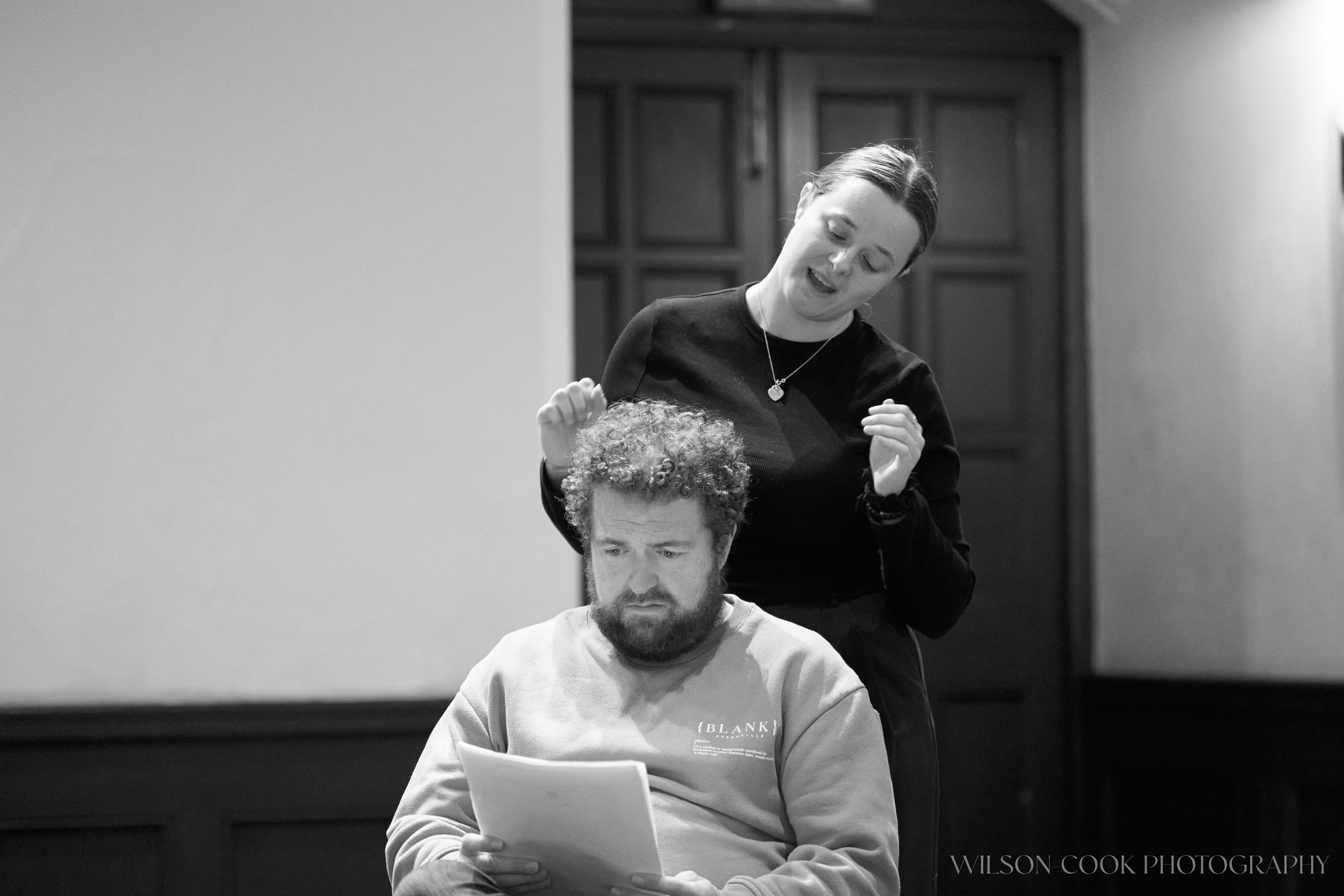 2025.  A black and white image of two actors rehearsing, a man and a woman.  The woman stands behind the man, as though she is admiring his hair.