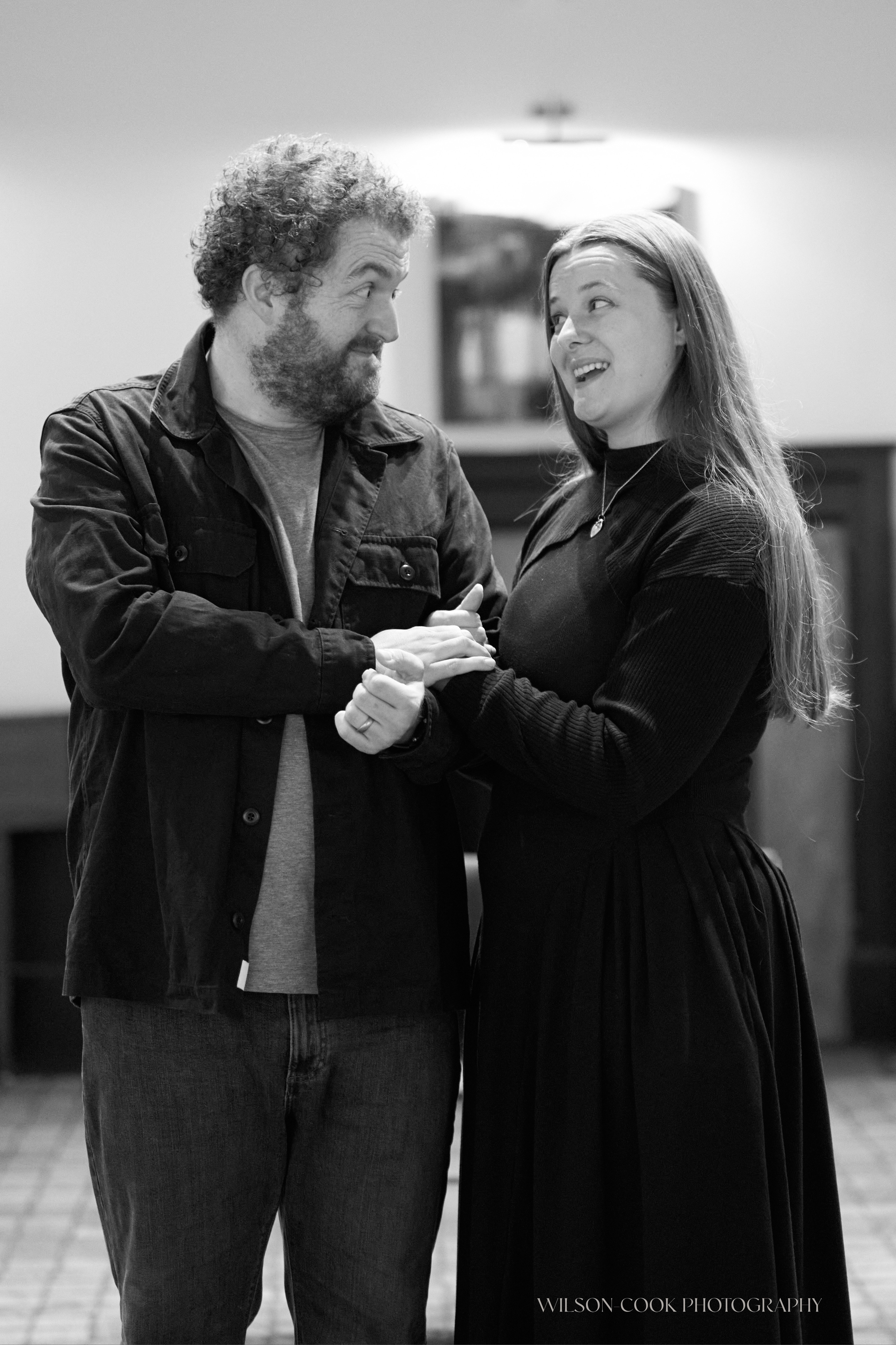 2025.  A black and white photo of two actors in rehearsal, a man and a woman.  The woman is holding onto the man's arm.