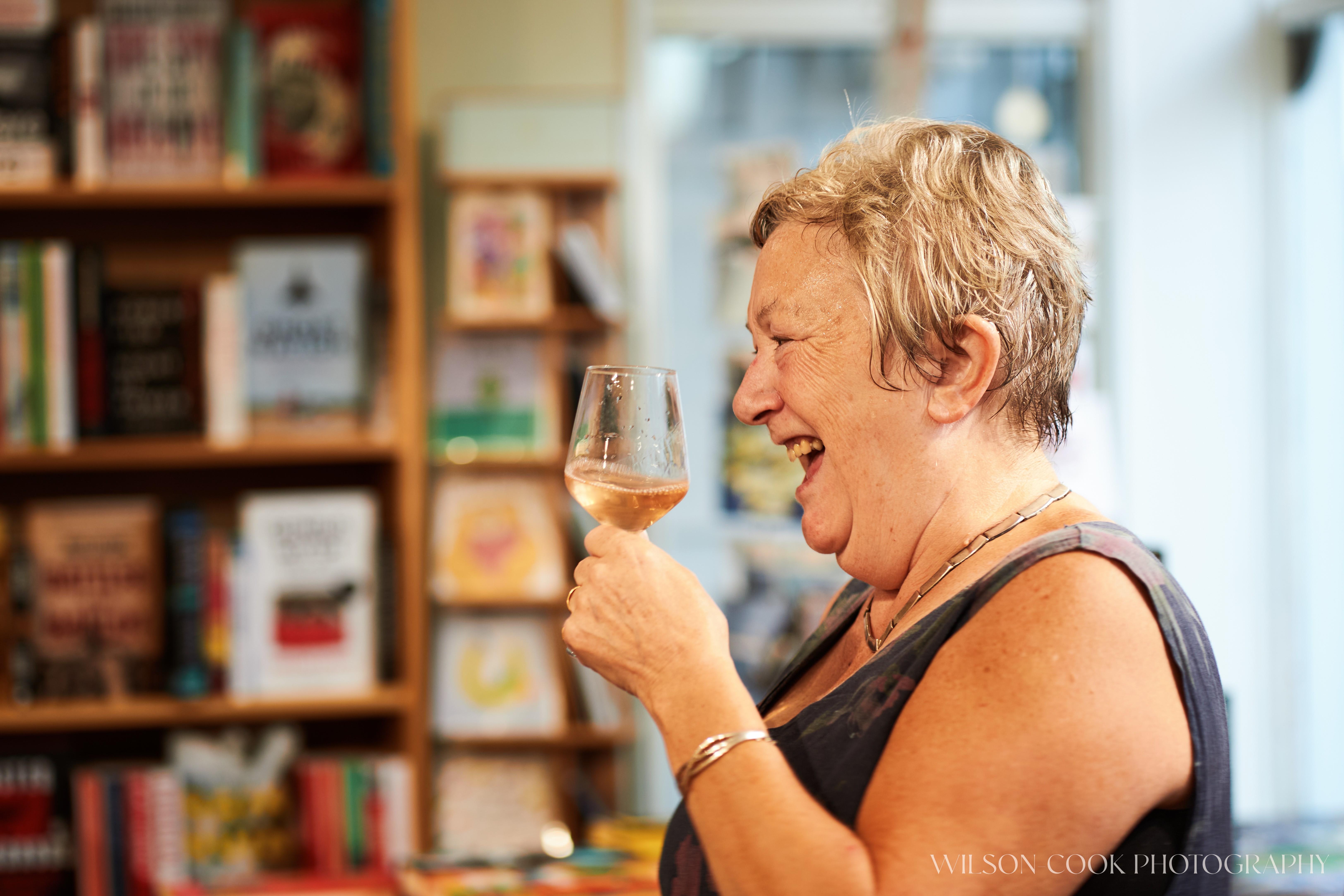 2025.  A woman stands, holding a glass of wine, facing the left of the frame, she is smiling as though she is mid-laugh.  Taken at Mostly Books, Abingdon.